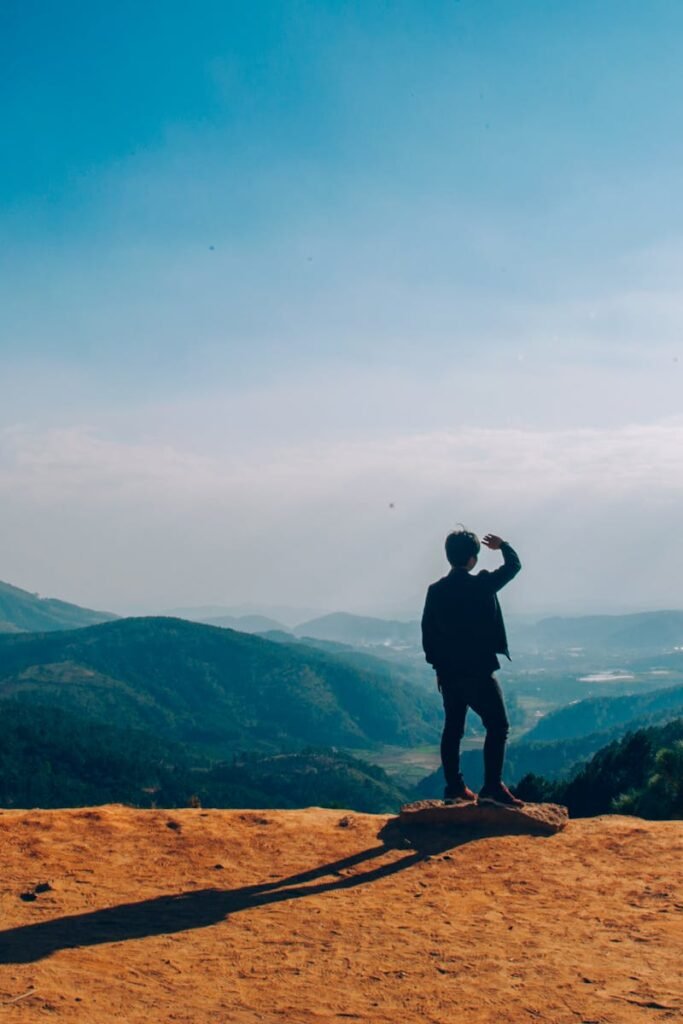 A lone traveler stands on a hilltop, gazing over a vast mountain landscape under a clear blue sky.